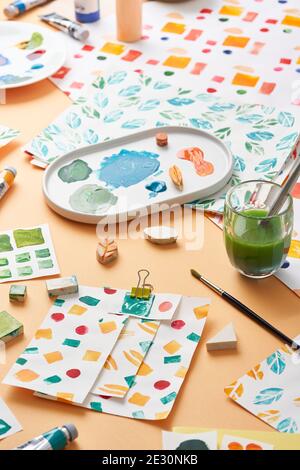 Platte mit Flecken von Farbe und Glas von farbigem Wasser Platziert auf orangefarbenem Hintergrund inmitten von Papierblättern mit verschiedenen Ornamenten Stockfoto