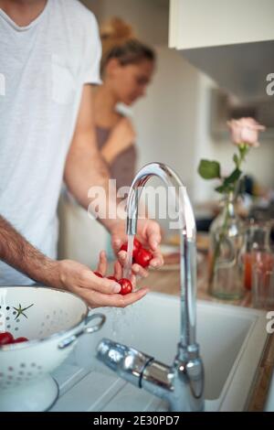 Ein junger Mann, der an einem schönen Morgen frische Kirschtomaten unter fließendem Wasser in der Küche wäscht. Haus, Küche, Salat Stockfoto