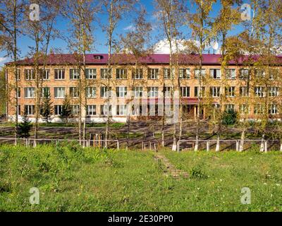 Die Fassade des Internats ist hinter Pappelbäumen an einem sonnigen Sommertag im Dorf Parnaya versteckt. Region Krasnojarsk. Russland. Stockfoto