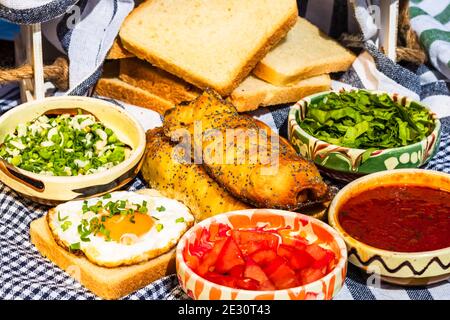 Rustikale Komposition mit Würstchenbrötchen, Spiegelei auf Toast, verschiedenen Schüsseln mit Sauce und gehacktem Gemüse. Stockfoto