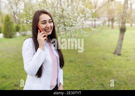 Ärztin spricht auf dem Handy. Medizinischer Hintergrund Kopie Raum. Im Freien eines Krankenhauses in Blumengarten. Fröhliche junge Frau. Stockfoto