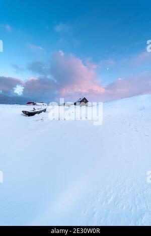 Holzboot und Holzhaus auf schneebedecktem Feld in Eine arktische Landschaft bei Sonnenuntergang mit dem Mond im Himmel Stockfoto