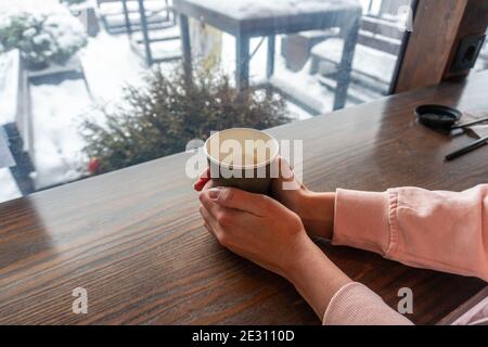 Ein Mädchen mit zwei Händen hält eine Papiertasse mit Kaffee in einem Café mit Blick auf eine Winterstraße Stockfoto