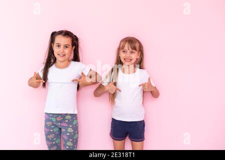 Staunende kleine Kinder Mädchen zeigt weißes T-Shirt, isoliert auf rosa Hintergrund. Lifestyle-Konzept für die Kindheit. Erstellen Sie einen Mock-up-Kopierbereich. Zeigefinger Stockfoto