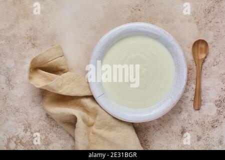 Griechischer Joghurt in weißer Schale, Leinenserviette und Holzlöffel auf altem beigen Tisch. Stockfoto