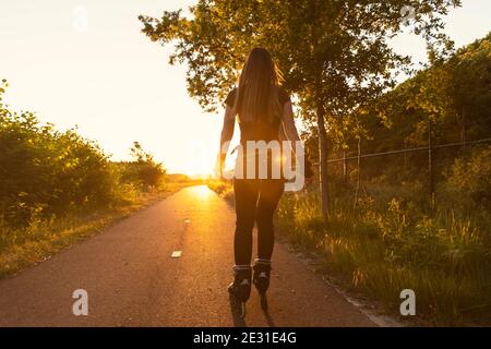 Eine junge Frau rollt auf einer Straße mit Grün im Hintergrund während des Sonnenuntergangs. Sportlicher, gesunder Lebensstil. Goldene Stunde und Sonneneruptionen Stockfoto