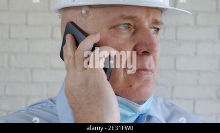 Ingenieur trägt Helm und schützende medizinische Gesichtsmaske sprechen Sie mit Smartphone in Coronavirus Pandemie Stockfoto