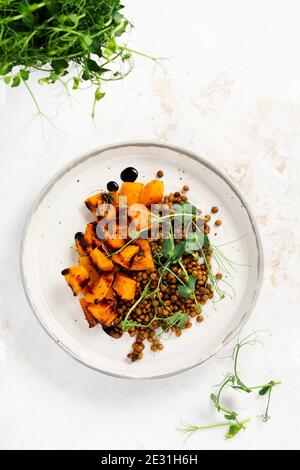 Salat mit gebackenem Kürbis, Linsen und Balsamico-Dressing garniert mit Erbsen-Microgreens auf hellem Hintergrund. Gesundes veganes Essen, Draufsicht. Stockfoto