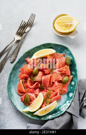 Scheiben leicht gesalzener Lachs mit Kapern, Zitrone und Dill auf einer Keramikplatte auf grauem Betongrund. Draufsicht. Stockfoto