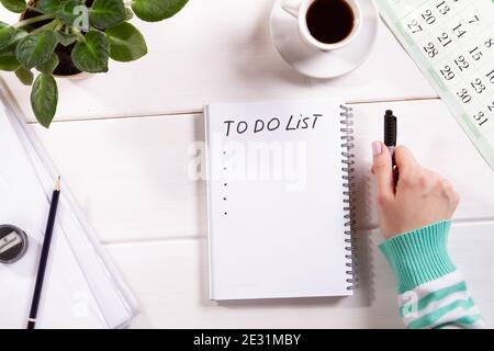 Draufsicht Mockup der Frauenhand mit Notizblock auf weißem Holz Hintergrund Kalender, Tasse Kaffee und Blume. Planung, To-Do-Liste oder Shopping. Entspannen Sie sich, w Stockfoto