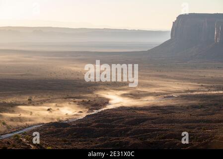 Sonnenlicht, das Staub auffängt, der von Fahrzeugen, die bei Sonnenaufgang über die Wüstenstraße fahren, im Monument Valley Navajo Tribal Park, Arizona und Utah, USA, aufgeworfen wird Stockfoto