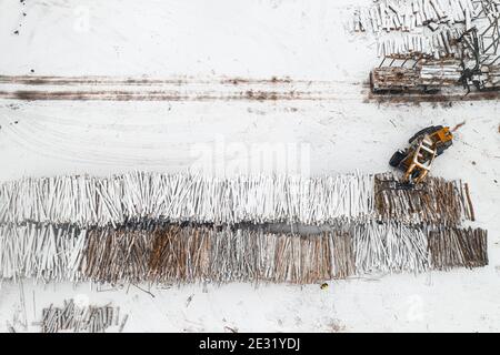 Der Lader lädt die in mit Schneedecke bedeckten Pfähle gestapelten Holzstämme Anzeigen Stockfoto