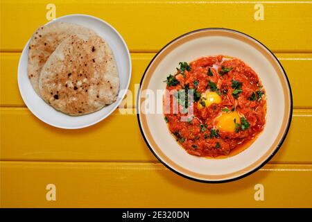 Shakshuka- und Pita-Brot - typisch israelisches Gericht mit Eiern mit Tomatensauce, Paprika, Petersilie und Gewürzen - Draufsicht flach auf gelbem Hintergrund Stockfoto