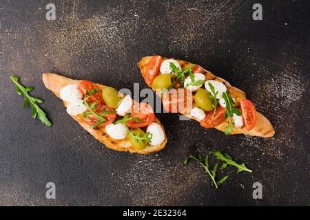 Caprese Bruschetta Toasts mit Kirschtomaten, Mozzarella, Oliven und Basilikum auf altem dunklen Hintergrund. Draufsicht. Stockfoto