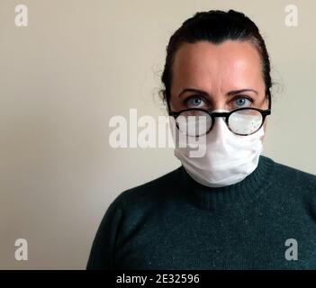 Frau in Brille mit weißer medizinischer Maske, Frau in weißer medizinischer Maske mit Brille mit Regentropfen, Frau in grünem Pullover vor Weiß stehend Stockfoto