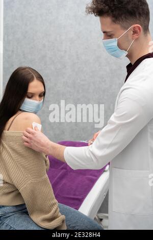 Ein Teenager sitzt in einer Arztpraxis und pickaxing für einen Impfstoff von einem jungen Arzt gegeben werden. Die COVID-Impfe19. Desinfiziert die abblätterende Schüssel. Stockfoto