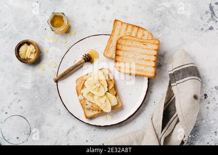 Toast mit Erdnussbutter, Bananenscheiben, Honig und Mandelflocken auf einem alten grauen Betongrund. Draufsicht. Stockfoto