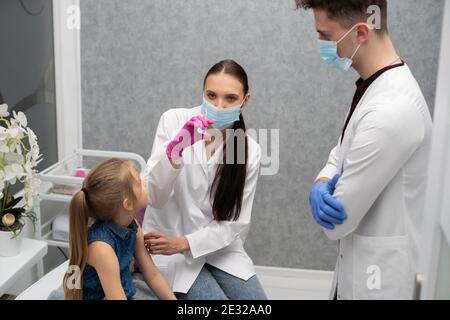 Ein Lehrling im letzten Jahr der Medizin spricht mit einem kleinen Patienten, um keine Angst vor Injektionen zu haben. Der Arzt bereitet sich auf die Injektion des neuen Impfstoffs vor. Stockfoto