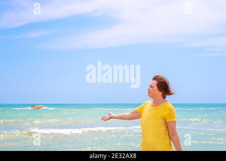 Eine ältere Frau an den Ufern des azurblauen Meeres gegen einen blauen Himmel, streckt ihre Hand zur Seite, und genießt einen Sommertag, das Ende der Quarantäne Stockfoto