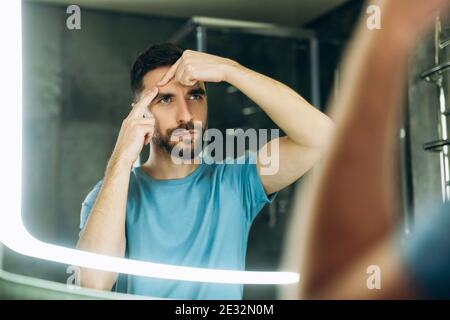 Junger Mann mit Bart in blau T-Shirt Quetschen Pickel im Badezimmer Spiegel Akne Problem. Stockfoto