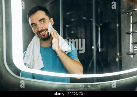 Nahaufnahme des attraktiven jungen Mannes mit Handtuch im Hals vor dem Spiegel stehend und Anwendung feuchtigkeitsspendende Creme auf sein Gesicht Badezimmer Morgen. Stockfoto