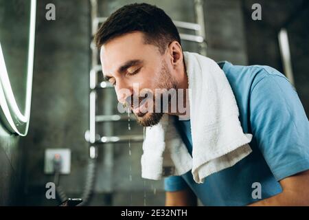 Junger Mann mit Bart waschen Gesicht. Person Reinigung Gesicht mit Handtuch auf Hals Wasch Seife im Waschbecken im Badezimmer zu Hause Stockfoto