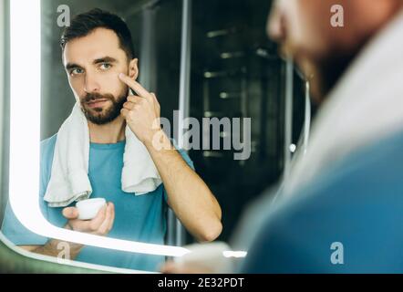 Nahaufnahme eines hübschen jungen Mannes mit einem Handtuch im Nacken, das vor dem Spiegel steht und feuchtigkeitsspendende Creme auf sein Gesicht auftragen. Stockfoto