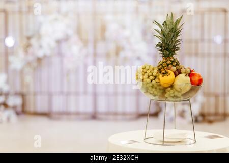 Glasschale mit Trauben, Ananas, Birnen, Pfirsichen und Äpfeln auf dem Metallständer auf dem unscharfen rosa Hintergrund mit dem Platz für Ihren Text Stockfoto