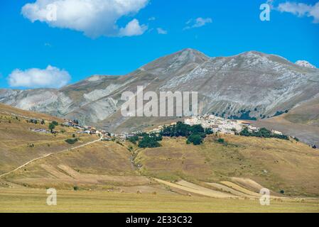 Die Stadt Castelluccio di Norcia von der Hochebene Pian Grande aus gesehen. Sommersaison, gelbes Gras. Castelluccio wurde vor kurzem von einem Erdbeben getroffen Stockfoto