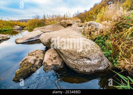 Ein schneller, flacher, sauberer Bach fließt zwischen glatten nassen großen Steinen, umgeben von hohen trockenen Klumpen, die im Wind im malerischen Ukraine schwanken Stockfoto