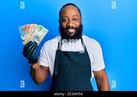 Junger afroamerikanischer Mann, der eine professionelle Schürze mit australischen Dollar trägt Banknoten, die positiv und glücklich stehen und lächeln mit einem Konfi Stockfoto