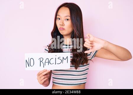 Junge schöne chinesische Mädchen hält keine Rassismus Banner mit wütenden Gesicht, negative Zeichen zeigt Abneigung mit Daumen nach unten, Ablehnung Konzept Stockfoto