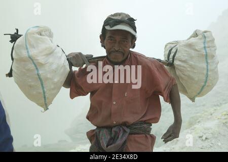 Miner Pak Agus, der älteste Schwefelminer, trägt die Säcke mit Schwefel aus den Schwefelminen im Krater des aktiven Vulkans Kawah Ijen in Ost-Java, Indonesien. Pak Agus ist jetzt 60 Jahre alt. Er arbeitet seit mehr als 40 Jahren in den Schwefelminen. Als er jünger war, konnte er mehr als 100 Kilogramm Schwefel vom Kraterboden nach oben tragen. Jetzt ist sein Limit 80 Kilogramm. Pak Agus ist der einzige arbeitende Bergmann seines Alters und er hat das Glück, noch am Leben zu sein. Die meisten Bergleute sterben im Alter von etwa 40 Jahren. Jeden Morgen klettern zwei Hunderte Bergleute auf den Grund des Kraters für die Stockfoto