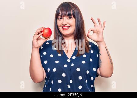 Junge plus size Frau hält roten Apfel tun ok Zeichen mit Fingern, lächelnd freundlich gestikulieren ausgezeichnete Symbol Stockfoto