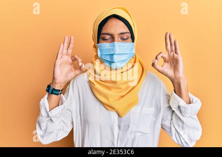 Junge Brünette arabische Frau tragen traditionelle islamische Hijab und medizinische Maske entspannen und lächeln mit geschlossenen Augen dabei Meditation Geste mit Fingern. Stockfoto