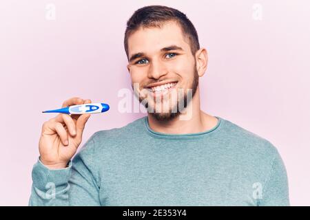 Junger schöner Mann hält Thermometer positiv und glücklich stehend Und lächelt mit einem selbstbewussten Lächeln, das Zähne zeigt Stockfoto