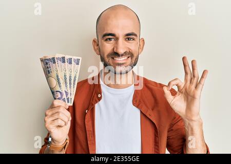 Junger hispanischer Mann hält 5000 japanische Yen Banknoten tun ok Zeichen mit Fingern, lächelnd freundlich gestikulieren ausgezeichnetes Symbol Stockfoto