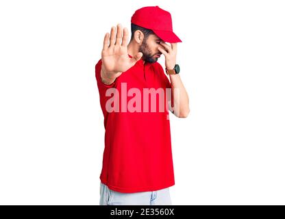 Junge gutaussehende Mann mit Bart tragen Lieferung Uniform Abdeckung Augen mit Händen und tun Stop-Geste mit traurig und Angst Ausdruck. Verlegen und n Stockfoto