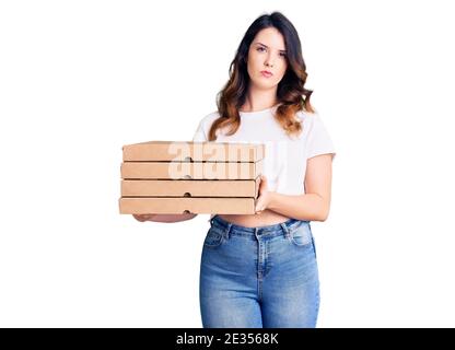 Schöne junge Brünette Frau mit Lieferung Pizza Box Denken Haltung Und nüchterner Ausdruck, der selbstbewusst aussieht Stockfoto