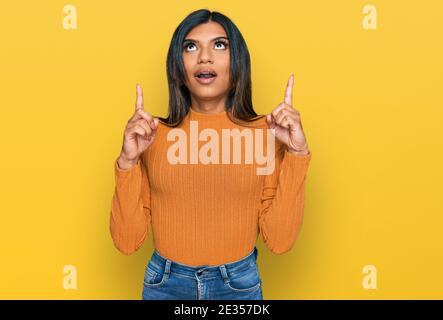Junge lateinische transsexuelle Transgender-Frau in lässiger Kleidung erstaunt und überrascht Blick nach oben und zeigt mit Fingern und erhobenen Armen. Stockfoto