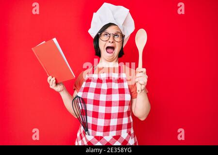 Brunette Frau mit Down-Syndrom tragen professionelle Bäcker Schürze lesen Kochen Rezept Buch wütend und verrückt schreien frustriert und wütend, shoutin Stockfoto