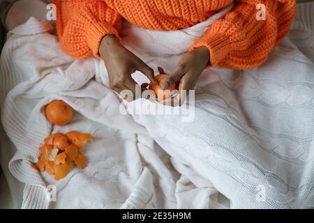 Afro Frau Hände schälen reife süße Mandarine, tragen orange Pullover, liegen im Bett unter dem Plaid Stockfoto