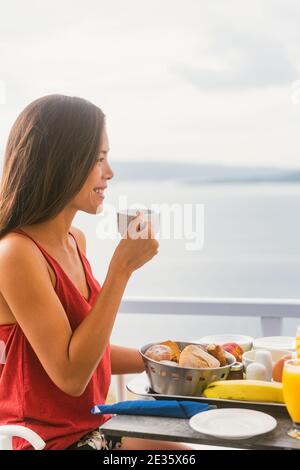 Kaffee trinken glückliche asiatische Frau trinkt Tee Tasse am Morgen Frühstück draußen in der Sonne genießen Essen im Hotelrestaurant oder Kreuzfahrtschiff Balkon Suite. Stockfoto