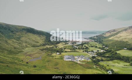 Dorf an grünen Bergtal Antenne. Schottische Stadt am Hang mit See, Burgruinen. Ländliche niemand Natur Landschaft. Whiskey Destillerie in Lochranza Countryside, Arran Island, Schottland, Europa Stockfoto