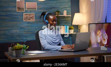 Porträt von glücklich lächelnden schwarzen Designer Frau mit Kopfhörern sitzen zu Hause Schreibtisch, arbeiten im Loft-Innenraum Überstunden Musik hören. Afrikanischer Freiberufler, der neues Projekt erstellt, das spät arbeitet Stockfoto
