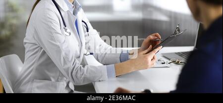 Unbekannte junge Ärztin hört ihrer Patientin zu, während sie in der Klinik zusammen sitzt. Medizinkonzept während der Coronavirus-Pandemie Stockfoto