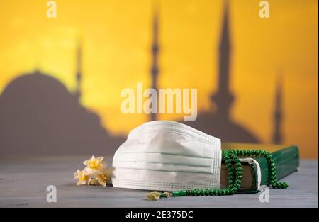 Koran, Rosenkranz und Schutzmaske auf dem Tisch. Moschee im Hintergrund bei Sonnenuntergang. Coronavirus Covid-19 Quarantäne muslimische Gebet Konzept. Stockfoto