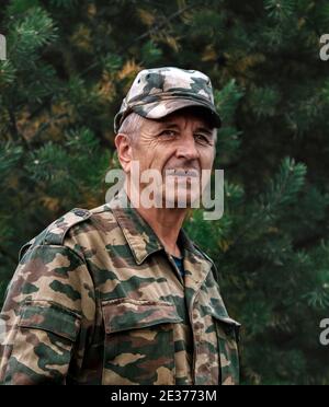 Älterer Mann in kunterbunten Jacke und Kepi gegen grüne Zweige der Kiefer. Selektiver Fokus Stockfoto
