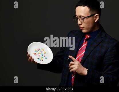 Junge asiatische Geschäftsmann Apotheker Arzt in karierten offiziellen Anzug, Krawatte und Brille hält zeigt Platte von medizinischen Pillen Stockfoto