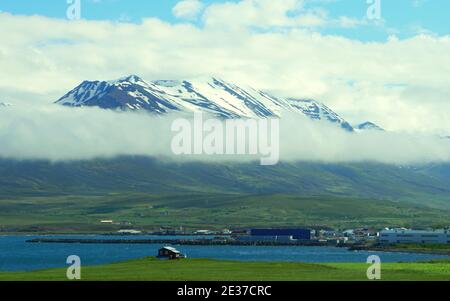 Die Fernansicht eines kleinen Hauses mit Blick auf den Schnee Und nebligen Berg und blauen Ozean im Sommer entlang Die Ringstraße in Island Stockfoto